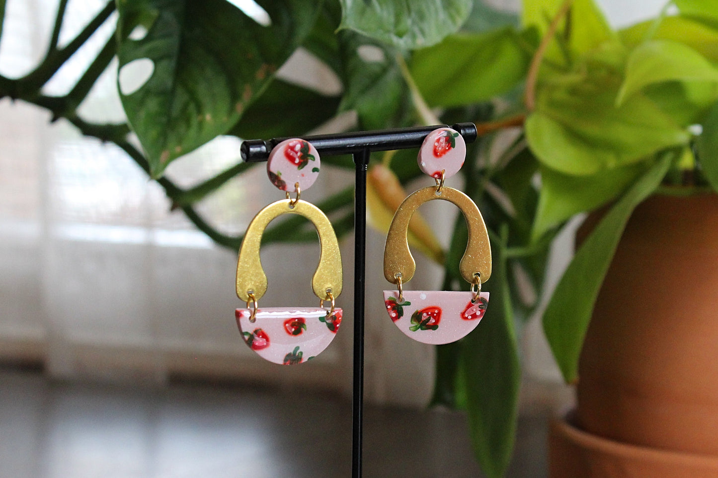 Hand painted strawberry drops earrings with gold arches and stainless steel posts displayed on black stand with green plants background