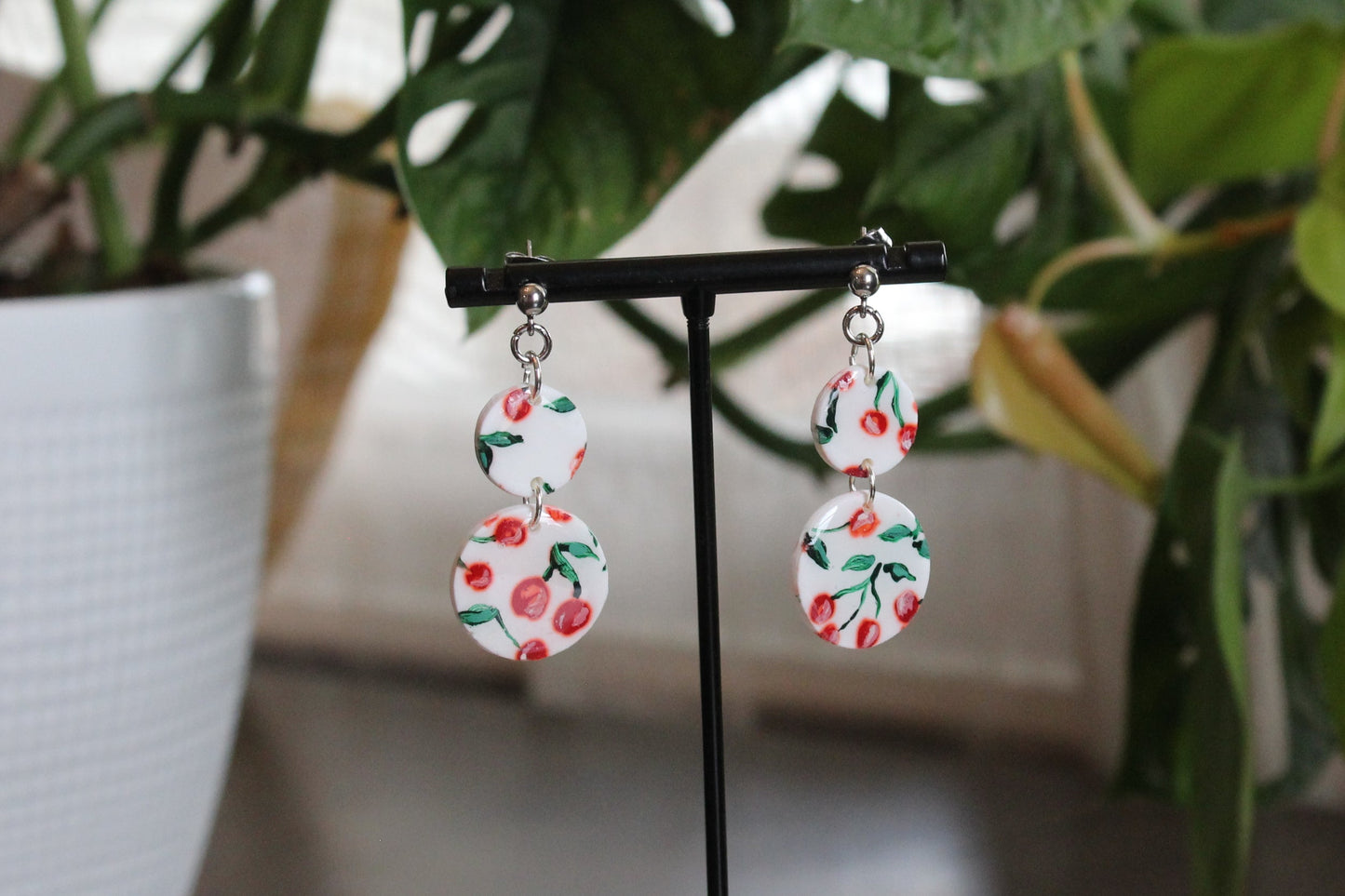 Hand painted cherry dangles on silver stainless steel ball studs with 1.75 inch drop hanging on a black display stand with green plants background