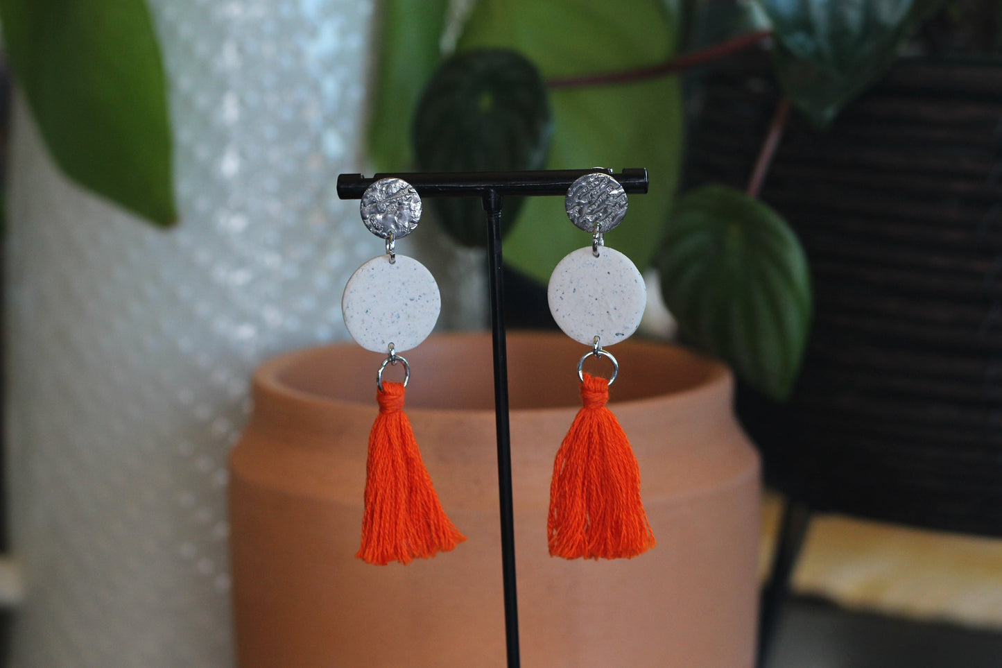 Speckled white tassel earrings with silver studs and orange tassels on display stand