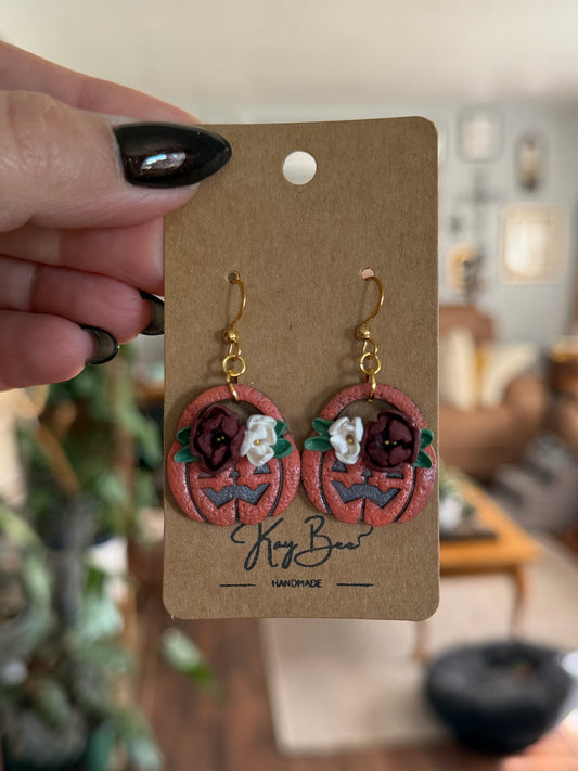 A hand with black nail polish holds a brown kraft card displaying a pair of handmade dangle earrings shaped like carved pumpkins. The earrings are terracotta orange with dark red and white details, gold hooks, and green leaf accents.