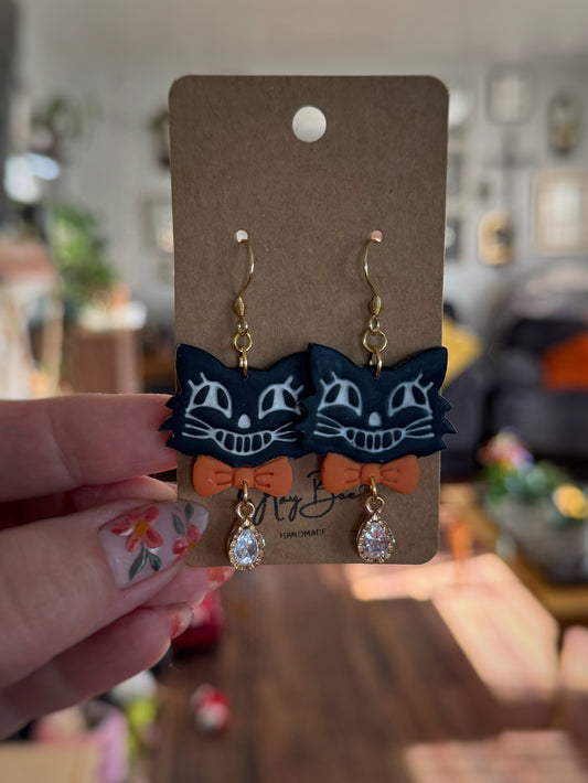 Pair of cat-shaped earrings with orange bows on a brown card held by a hand.