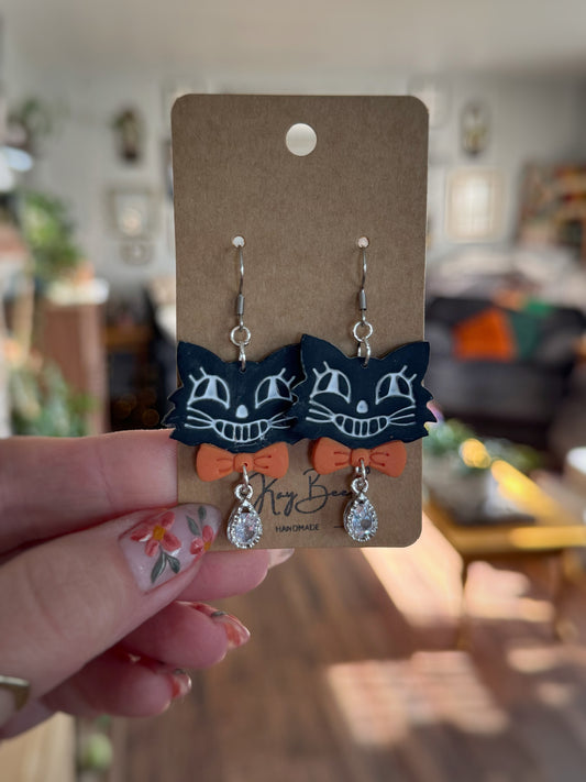 Cat-shaped earrings with orange bows on a cardboard backing held by a hand.